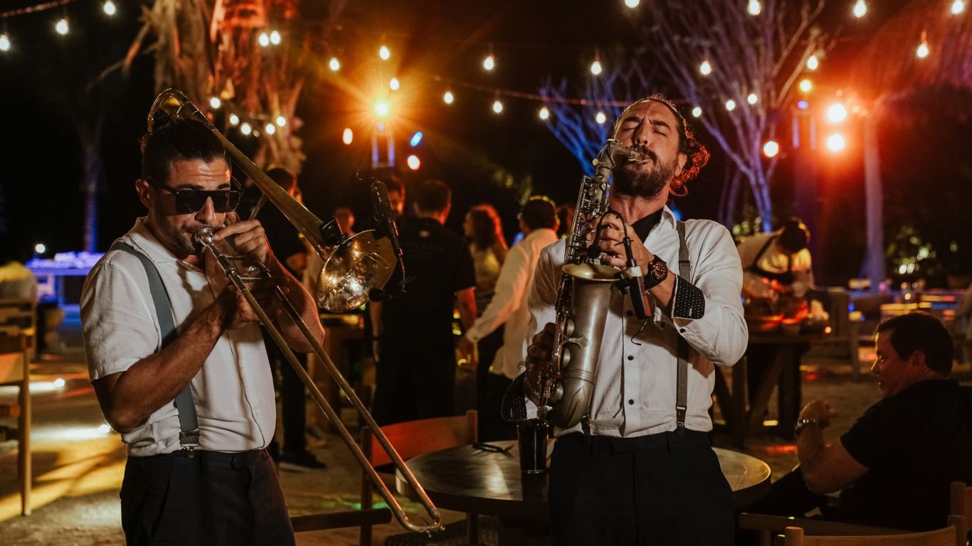  Dos músicos masculinos tocando el trombón y el saxofón afuera en un evento bajo las luces de Edison